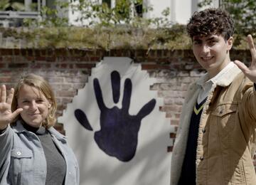 Auf dem Bild sind die zwei Hauptdarsteller:innen des Handabdruck-Films zu sehen, wie sie ihre Hände in die Kamera halten. Hinter ihnen ist ein Handabdruck auf der Mauer zu sehen.